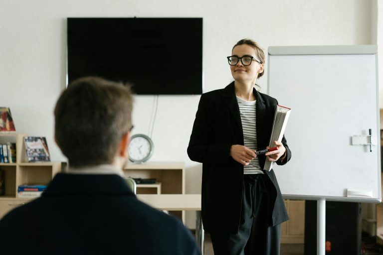 Caucasian female teacher in black blazer holding books, presenting to a student in a classroom.