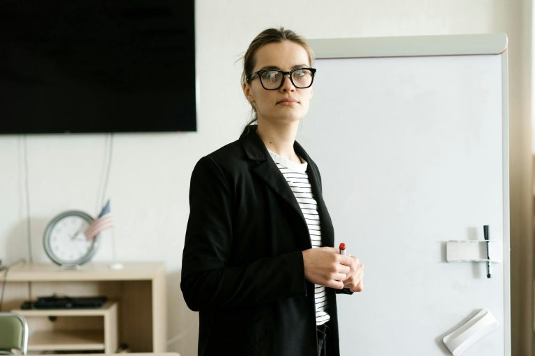 Caucasian woman instructor with glasses stands confidently by whiteboard in classroom.